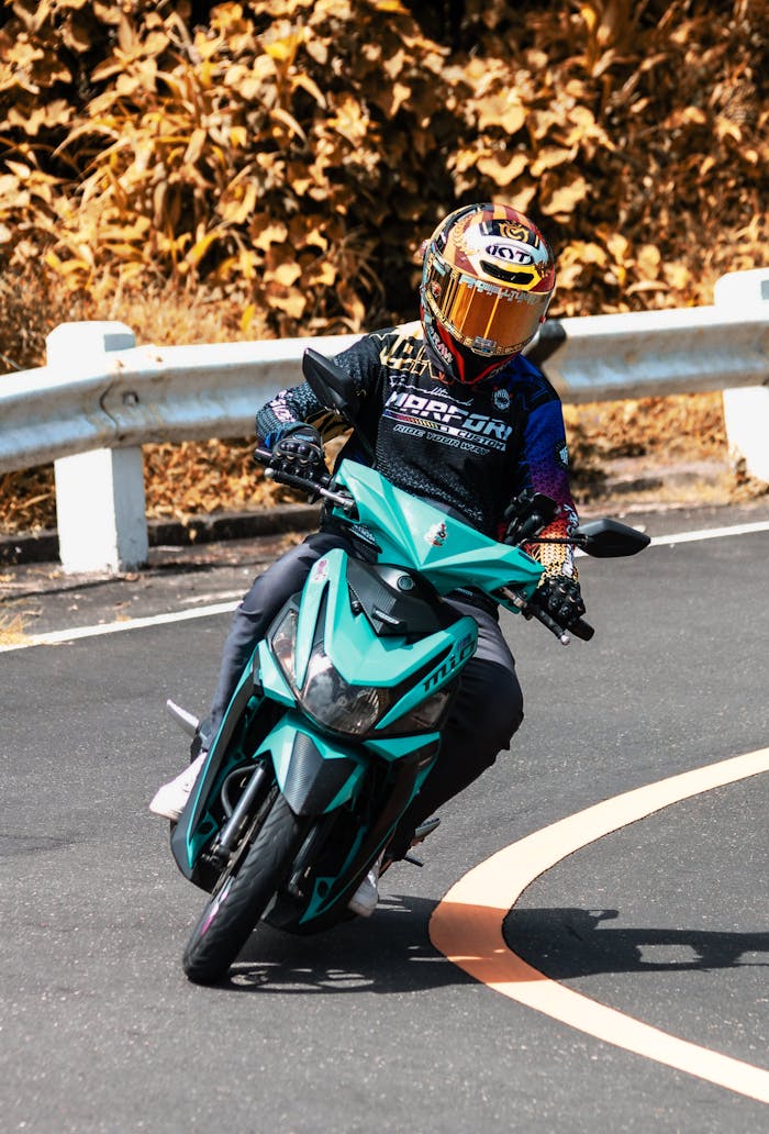 A motorcyclist in full gear maneuvers a teal Yamaha Mio through a city street curve. Safety gear is visible.